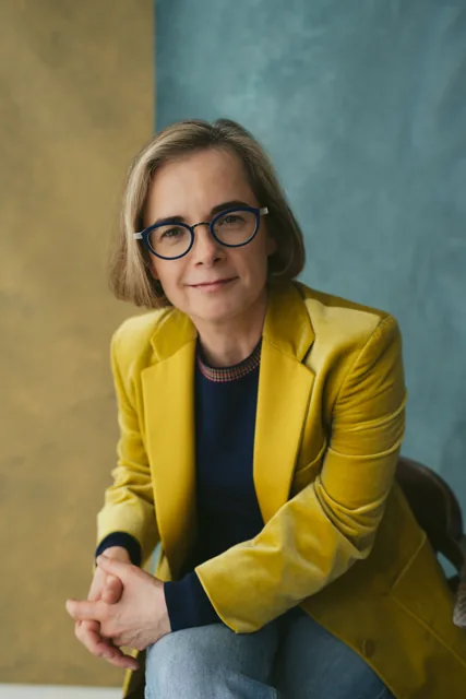 A woman in a mustard blazer against a blue background.