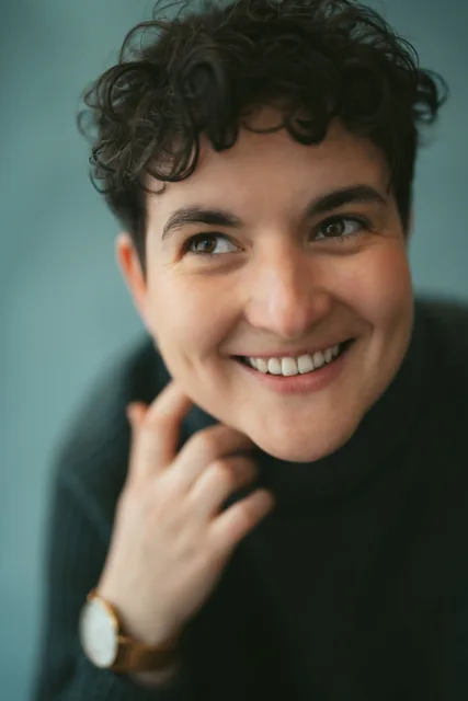 A smiling sitter with dark curly hair against a blue-green background.