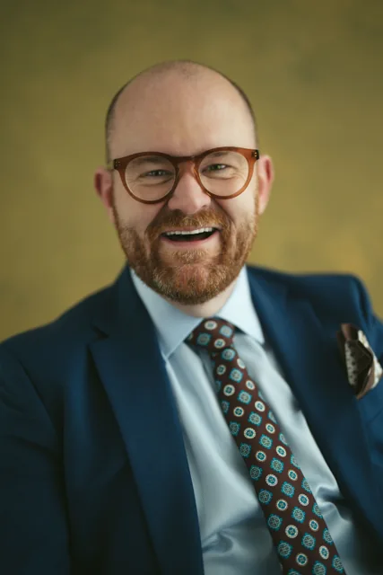 A laughing man in a navy suit against a mustard background.