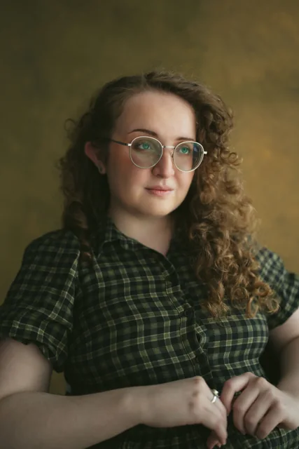 A curly-haired sitter with round glasses against an olive background.